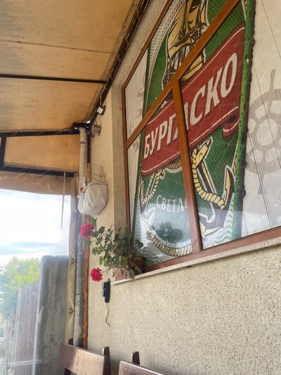 The image captures a detailed view of a semi-enclosed outdoor seating area located in Konstantinovo, Bulgaria. The primary visual element is a light-colored, granular-textured wall on the right, which features a large rectangular window-like structure framed in dark brown wood. This structure encloses a translucent screen displaying a large graphic advertisement for "БУРГАСКО" (Burgasko) beer. The advertisement design includes a golden sailing ship, a large anchor on a green background, and simulated water droplets. Below the graphic, the word "СВЕТА" is partially visible.
A small potted plant with vibrant red flowers rests on the windowsill ledge. Below the window, a black, two-gang electrical switch or outlet is mounted on the wall, with an exposed wire trailing downwards. Partially visible at the bottom of the frame are the dark brown wooden backs of what appear to be benches or chairs.
To the left of the window structure, a vertical, light-colored utility pipe or drainpipe descends along the wall. Attached to the pipe and the wall are various wires and a white, fabric-like bag or container suspended by a wire grid. Above this, a light-colored canvas or fabric canopy forms the ceiling, supported by dark metal beams. Exposed electrical wiring runs along these beams, leading to small, dark spotlight fixtures.
The left side of the image reveals an exterior view through a translucent, clear plastic or tarpaulin curtain. This view shows an overcast sky, green trees in the distance, and a prominent, rough-textured concrete wall or fence structure closer to the foreground. No individuals are present in the scene, and no specific actions are occurring.