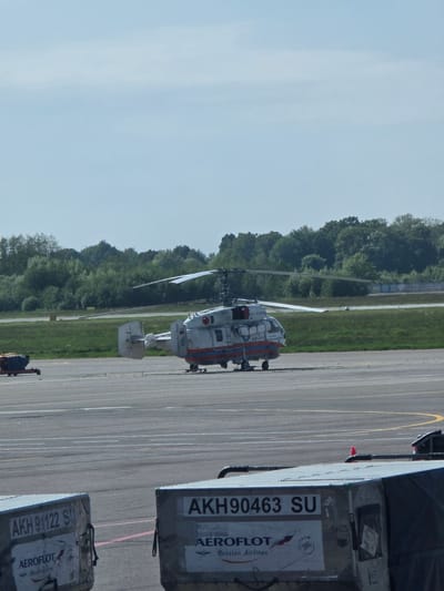 A helicopter is positioned on an airfield runway adjacent to a grassy area and a line of trees under a clear sky. There is cargo next to it marked with Aeroflot Russian Airlines, and the alphanumeric code AKH90463 SU. The location is Khrabrovo, Russia. A person is seen near the bottom of the image. The scene appears to be taken from within a plane.