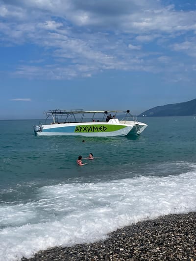 In Gagra, Abkhazia, on a bright, sunny day with scattered clouds, the scene unfolds at the edge of a calm, turquoise sea meeting a pebble beach. A modern, white and green catamaran, labeled with the Cyrillic word "АРХИМЕД," floats gently in the shallow water, its canopy providing shade for several passengers. Two individuals are swimming near the shore, their heads visible above the water, while another person's head is submerged. Waves are lapping at the beach, creating a frothy white foam that gently rolls onto the shore, revealing a colorful array of dark pebbles. In the background, a verdant, mountainous coastline stretches across the horizon, with the silhouette of a town faintly visible on its slopes.