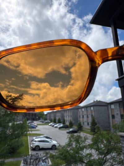 A close-up shot of sunglasses with amber-tinted lenses, held up to the sky, frames a reflection of clouds and a tree. In the background, out of focus, is a parking lot with several cars, a row of apartment buildings, and lush green trees. The scene is set against a backdrop of a partially cloudy blue sky. The architecture of the buildings suggests an urban or suburban environment. The presence of parked cars and apartment buildings indicates this is likely a residential area. The overall setting suggests a temperate climate, given the greenery and the use of sunglasses. While the specific location in Gatineau, Canada cannot be definitively determined from the image alone, the presence of apartment buildings and parking lots is common in many urban and suburban areas.