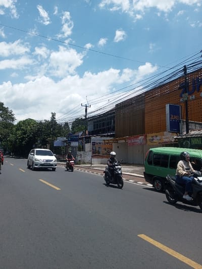 This image captures a daytime street scene in the city of Tagtse Tashidingkha, Bhutan, under a blue sky with scattered white clouds. The asphalt road, marked with yellow lane lines, shows traffic moving away from the viewer. From left to right, a partial red vehicle, a white Toyota Innova, a person on a red scooter wearing a dark helmet, and a person on a black scooter wearing a white helmet are visible. Further right, a green minivan travels alongside a person riding a dark scooter, who wears a black helmet, a light-colored long-sleeved top, and blue jeans. The right side of the street features a sidewalk bordered by commercial buildings. A prominent orange building with a perforated facade displays a large blue 'ZUKO' sign and a smaller 'ZUKO SHOP' sign. Adjacent buildings exhibit dark windows and some wooden paneling. Banners, including a red one displaying 'BUNGA' and another referencing 'NASI', are affixed to the storefronts. Overhead utility wires traverse the scene, supported by a visible power pole. Trees line the left side of the street and appear in the background, indicating urban vegetation.