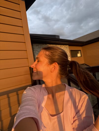 In Gatineau, Canada, a woman with brown hair tied in a ponytail is seen from the chest up, looking to her left. She is wearing a light pink, textured cardigan over a matching top. The setting appears to be an outdoor balcony or deck, with wooden siding of a building visible on the left and the roofline of another building in the background. The sky is cloudy. The scene is bathed in warm, golden hour sunlight.