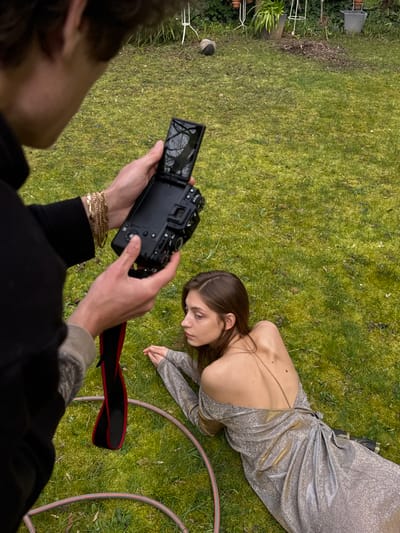 An outdoor scene in a garden or backyard within Vincennes, France, features two individuals. In the foreground, an individual, partially visible from the chest up, holds a black digital camera with both hands, angled towards a second subject. The camera's flip-out screen is open and displays a reflection of bare tree branches. The individual operating the camera wears a dark long-sleeved top, with multiple gold-colored chain bracelets on the left wrist, and a black camera strap with a red underside hangs from the device.
The second individual, a female, lies prone on green grass in the middle ground, facing left with her head turned slightly away from the camera's direct line of sight. She is positioned on her stomach, with her hands clasped in front of her. She wears a shimmering, silver-toned, long-sleeved dress with an off-the-shoulder design and a deeply cut open back, exposing her upper back and shoulders. A thin, light-colored strap is visible on her upper back. Her hair is dark brown and parted on the side. A grey garden hose is coiled on the grass, partially encircling her lower body.
The background consists of a well-maintained lawn with scattered grass clippings, behind which are various garden elements including white metal plant stands, terra cotta pots, a grey bucket, and some green foliage from potted plants. A brown object rests on the grass further back. The scene depicts a photographic or videographic session in progress.