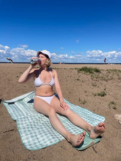 An overhead medium shot shows a woman sitting on a beach blanket on a sandy beach. The woman is wearing a white bikini and a white bucket hat. She is holding a can and drinking from it with her right hand. Her legs are extended out in front of her. The blanket is green and white checkered. The background shows a beach with a few people in the distance, a body of water, and a blue sky with white clouds. The scene appears to be taking place on a sunny day. There is no identifiable landmark linking this image to the city of Belozersk, Russia.