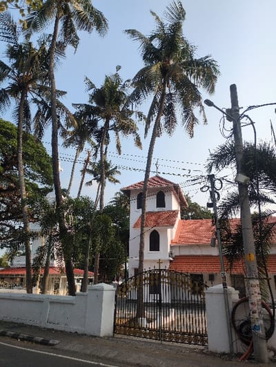 This image depicts the St. Francis CSI Church, located in Fort Kochi, Kochi, Kerala, India, viewed from a roadside perspective on a clear day. The primary subject is a multi-story white building with a prominent red-tiled roof. A distinctive white square tower with a red pyramidal roof and dark, arched windows rises from the structure, slightly right of center. The church's white walls contrast with its red clay tile roof sections. A small cross is affixed to the church facade just above the main gate. In the background, to the left of the main church structure, another white, dome-roofed building is partially visible amidst dense green foliage.
In the foreground, a low, white concrete compound wall separates the church property from a paved road, which features a black-and-white striped curb marking. A black wrought-iron gate with intricate gold decorative elements forms the main entrance through the wall.
Numerous tall, slender palm trees with green fronds dominate the upper and mid-sections of the image, partially obscuring the church building and extending into the clear blue sky. Additional green trees and shrubbery are visible within the church compound.
On the far right, a grey utility pole stands upright, supporting a black street lamp fixture that extends to the left. Multiple black electrical wires crisscross the sky from this pole. A bundled black cable is attached to the lower section of the pole, which also displays a yellow circular sticker marked with a 'T'. No people or vehicles are present in the scene.