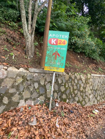 A green and yellow sign with white lettering is mounted on a metal pole, attached to a concrete post. The sign reads "Apotek K24" with an arrow pointing upwards. The sign is partially obscured by a tree trunk. The sign is located on a dirt slope with a stone wall. There are brown leaves on the ground. The location is on a street in Lembang, Indonesia, at the coordinates -6.828, 107.607. The sign is likely for a local pharmacy.