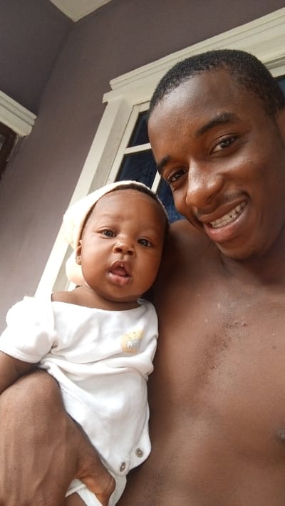 A man and a baby are captured in a close-up, brightly lit portrait, likely taken in Agburu Uke, Nigeria. The adult man, positioned on the right, has dark skin, short dark hair, and is shirtless. He is smiling broadly, revealing braces on his upper teeth, and appears to be holding the baby.
The baby, located on the left side of the frame, also has dark skin and is wearing a white long-sleeved onesie with snap closures at the crotch. A small, faded light yellow animal design, possibly a chick or duckling, is visible on the upper left chest of the onesie. The baby is also wearing a light-colored, possibly off-white, hat or head covering, and a small earring can be seen in their visible ear. The baby's expression is neutral and curious, with wide eyes looking directly at the camera and a slightly open mouth.
The background shows a grey wall and white architectural trim, possibly around a window or door frame of a building, suggesting the photo was taken outdoors or in a semi-sheltered area. The natural, soft lighting indicates it is daytime. The overall mood of the image is one of warmth and affection, highlighted by the man's happy expression. There is no visible text in the image.