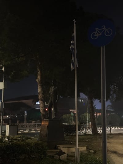The vertical image, captured at night, presents a somber outdoor scene in Egkomi Lefkosias, Cyprus. The dominant subjects are a statue, a flagpole bearing a flag, and a bicycle path sign, all set against a backdrop of dark trees and dimly lit structures.

In the foreground, a dark, full-body statue of a male figure, possibly a historical or military personality, stands on a dark, rectangular pedestal. A faint red light is visible near the base of the statue. On the pedestal, a light-colored plaque with unreadable text is affixed.

To the left of the statue, a flagpole extends upwards, displaying what appears to be the national flag of Greece. The flag, featuring nine horizontal blue and white stripes with a white cross on a blue canton, is partially furled. Above the flag and flagpole, a circular blue sign with a white bicycle symbol indicates a designated bicycle route or area.

The setting is characterized by lush, dark green foliage and trees that fill much of the background and mid-ground, suggesting a park or a green public space. In the distance, faint artificial lights from buildings illuminate parts of the dark sky. A metal fence and indistinct white road markings or a patterned wall are vaguely discernible in the background, hinting at an adjacent road or built environment. The overall mood is quiet and still, typical of a nighttime urban or suburban landscape.