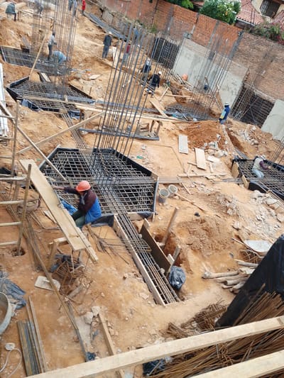 The image depicts an active construction site in Santa Cruz de la Sierra, Bolivia. The scene is characterized by an uneven, reddish-brown dirt terrain with numerous steel rebar structures, wooden formwork, and piles of construction materials. In the foreground, a construction worker wearing an orange hard hat, a dark jacket, and a blue shirt is positioned on a wooden plank, seemingly working on the rebar. To the left, a ladder and other scaffolding elements are visible. Further into the scene, other workers are engaged in various construction tasks, including one kneeling and working on a rebar grid, and another in the background near a brick wall. The environment shows signs of ongoing foundation work for a building, with concrete poured into some sections and rebar cages ready for concrete. The background reveals unfinished brick walls and vegetation, indicating a developed urban or semi-urban area within Santa Cruz. The overall impression is one of diligent manual labor on a construction project.