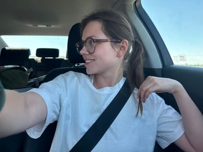 A young woman with brown hair and glasses is sitting in the passenger seat of a car, looking out the window to her right and smiling. She is wearing a white t-shirt and a black seatbelt. The interior of the car is visible, including the headrests and a portion of the dashboard. The background is a bright, clear sky. The image appears to be taken in Magnuszowice, Poland, a rural area.