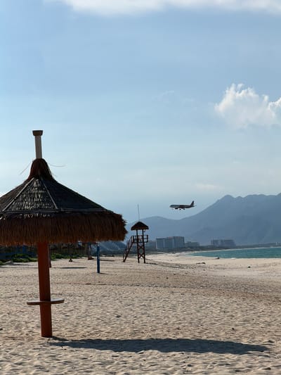 A beach umbrella with a thatched roof stands on a sandy beach. In the background, a lifeguard tower is visible, along with buildings and mountains. An airplane is flying in the sky above the mountains. The image appears to be taken in the city of Bac Cam Ranh Ward, Vietnam.