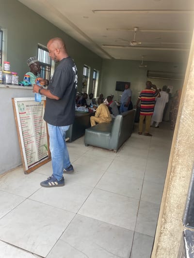 The image depicts an indoor scene that appears to be a waiting area or reception of a natural therapy clinic in Bauchi, Nigeria. In the foreground, a bald man wearing a black t-shirt and blue jeans is holding a plastic bottle of water. He is standing next to a wooden board with a list of ailments and treatments written on it, titled "IBRAHIM BABA JIMOH, N.D. NATURAL THERAPY CONSULTANT." The board is positioned on an easel.
In the background, several other individuals are visible. To the left, a man wearing glasses and a cap is behind a counter, interacting with the man in the foreground. Further back, there are people sitting on a green sofa and standing. One man in a striped shirt is on his phone. The room has tiled floors and a white ceiling with ceiling fans. A television is mounted on the wall. The overall environment suggests a functional, albeit somewhat dated, establishment. The natural light filtering in from the left suggests the entrance or a window is in that direction.