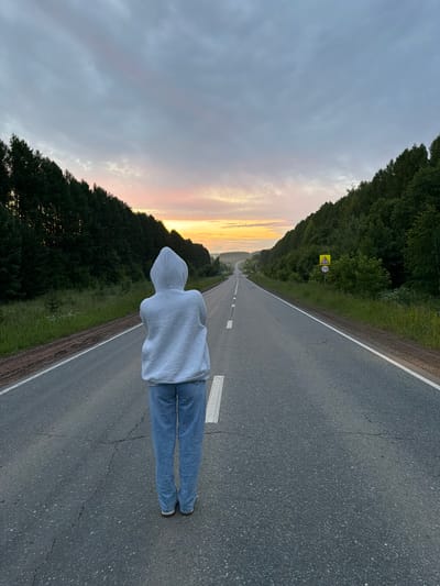 The image shows a person wearing a gray hoodie and blue jeans standing on an asphalt road, facing away from the camera. The road stretches into the distance, flanked by dense green trees on either side. The sky is overcast with hints of a sunset in the distance, casting a soft, diffused light over the scene. There are road markings and a speed limit sign visible on the right side of the road. The overall setting appears to be a rural or semi-rural area.