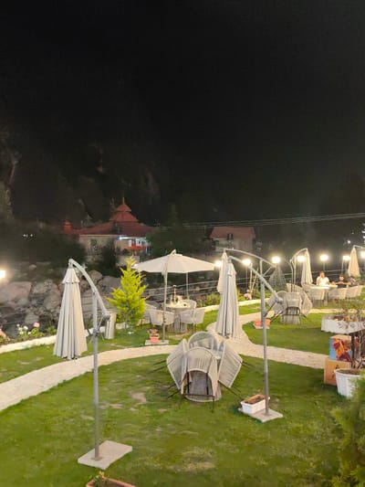 The image depicts an outdoor seating area at night, likely a restaurant or cafe, situated near a mountainous landscape and possibly a body of water. In the background, a building with a red, tiered roof, resembling a temple or traditional Indian architecture, is visible. The foreground is dominated by a well-maintained lawn with several white patio umbrellas and a curved, light-colored pathway. Outdoor furniture, including chairs and tables, is arranged on the lawn and pathway. Some chairs are stacked. Several lampposts with bright lights illuminate the area. In the mid-ground, a small, bright green evergreen tree stands near a cluster of tables. Two individuals are seated at one of the tables in the mid-ground, appearing to be engaged in conversation. The overall impression is a serene and inviting atmosphere for evening dining or relaxation in Kulang, India.