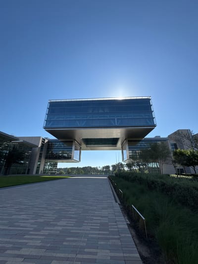The image shows a modern building with glass and stone or concrete facade in Spring, Texas, USA. The building has an angular, cantilevered design, with a large glass section appearing to float above a lower section. The sky is clear and bright blue. A paved pathway or walkway, made of gray and white bricks, extends away from the viewer alongside the building. A strip of green grass separates the walkway from the building and some greenery like bushes and trees. Sunlight reflects off the glass of the building.