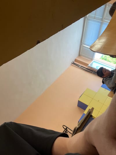 The image is taken from a low angle, looking upwards in a room that appears to be a school or educational facility. A person wearing a gray shirt is visible looking down in the distance. A table is in the foreground, and the room contains a window with blinds. The walls are peach colored. There are yellow square objects. A person with a black shirt appears to be taking the picture. This location is probably not in the city of Kavarna, Bulgaria.