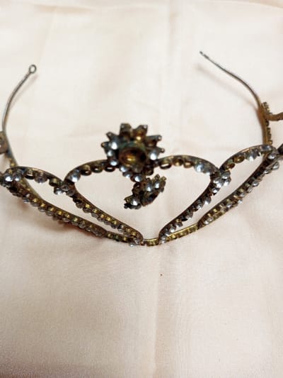 A tarnished metal tiara, or headband, adorned with small clear stones is centrally positioned on a light beige, possibly fabric, surface exhibiting soft folds. The tiara features an arched, openwork design composed primarily of interconnected heart-shaped segments, each lined with small, circular, clear stones. A prominent floral motif, also set with clear stones around a central larger, darker stone, is located at the apex of the tiara. Several stones are visibly missing from various settings, particularly from the central floral element and the adjacent heart segments. The metal frame displays discoloration consistent with age and oxidation, appearing brassy or bronze-toned. The two ends of the tiara are bent outwards and slightly upwards, suggesting a mechanism for securing it. The scene is located in Qarabucaq qəsəbə, Azerbaijan.