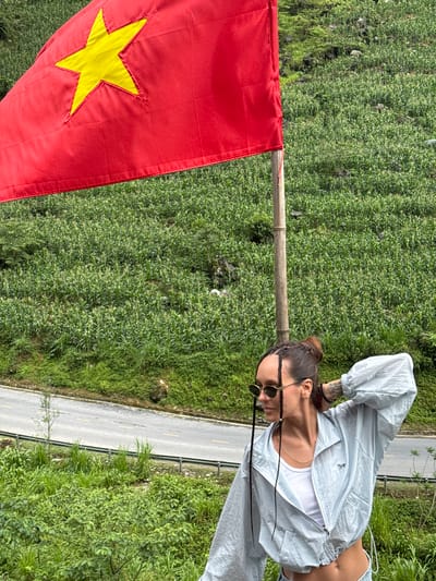 A vertical, medium shot captures a woman posing beside a Vietnamese flag against a backdrop of lush greenery in Yên Minh Commune, Vietnam. The flag, predominantly red with a central yellow star, is positioned on the left side of the frame. The woman stands on the right, wearing sunglasses, a white top, and a light blue jacket. A paved road runs along the edge of the greenery behind her. The dense vegetation, likely part of the landscape of Yên Minh, fills the majority of the background.
