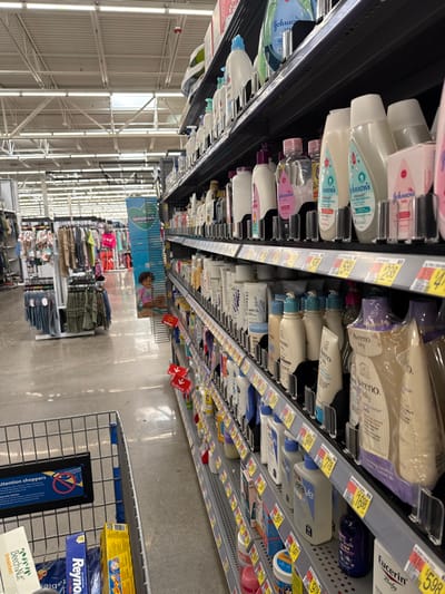 The image depicts a retail store aisle, likely a supermarket or large department store, focused on personal care or baby care products. The perspective is from eye level, looking down a long aisle between two rows of shelves. The image is rotated 90 degrees clockwise, so shelves appear on the left and right, and the floor runs horizontally in the lower part of the frame, with the ceiling in the upper part.

On the left side of the image, running vertically from top to bottom (if viewed upright), are multiple shelves stocked with various liquid products in plastic bottles. Many of these are baby care items, with "Johnson's" visible on several labels, including baby shampoo, lotion, and oil. The bottles are primarily white, pink, and light blue. Price tags are affixed to the shelf edges, displaying numbers like "208" and "144".

On the right side of the image, another set of shelves runs parallel to the first. These shelves also contain numerous personal care products. Brands like "Aveeno" (visible on a beige-colored package and white bottles), "Honest" (on white bottles with purple labels), "CeraVe" (on white and blue bottles), and "Eucerin" (on a purple bottle) can be identified. These shelves also have numerous price tags, with some visible prices like "$189", "$778", and "$598".

Further down the aisle, towards the background, there's a wider view of the store. On the left (if viewed upright), the store's ceiling structure with white trusses and lighting is prominent. On the right, beyond the shelves, there are racks of clothing, displaying folded items and hanging garments. A blue banner sign with an image of a child and the text "splast" (partially visible) is hanging above these clothing racks.

The floor of the aisle is a light gray concrete or polished surface. In the lower right foreground, a metal shopping cart is partially visible, with a blue handle. Attached to the front of the cart is a black sign with white text.

Visible text in the image includes:
*   "Johnson's" (on multiple baby product bottles)
*   "Aveeno" (on various bottles and packages)
*   "HONEST" (on white bottles)
*   "CeraVe" (on white and blue bottles)
*   "Eucerin" (on a purple bottle)
*   "splast" (on a blue banner in the background)
*   Price tags with numbers like "208", "144", "$189", "$778", "$598".
*   On the shopping cart sign: "attention shoppers", "Reyno", "Beech Nut", "breakfast".
*   "CNY" and "ables" are also visible on some product packaging near the cart.

The lighting appears bright and even, typical of a retail environment, suggesting it's daytime. No people are clearly visible in the immediate foreground or middle ground, but the presence of shopping carts and stocked shelves indicates an active store setting.