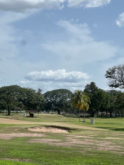 This is a landscape photograph of a golf course under a cloudy sky in Maracay, Venezuela. The foreground features a sand trap and green grass. In the mid-ground, there is a gently sloping fairway with a white marker post. Further back, a small pavilion with a brown roof and red walls stands near a flagpole on the putting green. Several mature trees, including large leafy ones and a palm tree, dot the landscape. In the far background, a white fence encloses what appears to be a stable or equestrian facility, with a colorful playground structure visible beyond it. The sky is partly cloudy with white cumulus clouds against a light blue background.