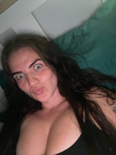 The image shows a close-up, high-angle shot of a woman lying down. She has long, dark brown hair and is looking directly at the camera with wide, light blue eyes. Her lips are puckered as if for a kiss, and her eyebrows are notably dark and well-defined. She appears to be wearing a black top, with her chest partially visible. The background consists of a green pillow and a white surface, possibly a headboard or wall. The lighting suggests an indoor environment, likely a bedroom. There is no indication of the location being in Мосметаллоконструкция Онт, Russia.