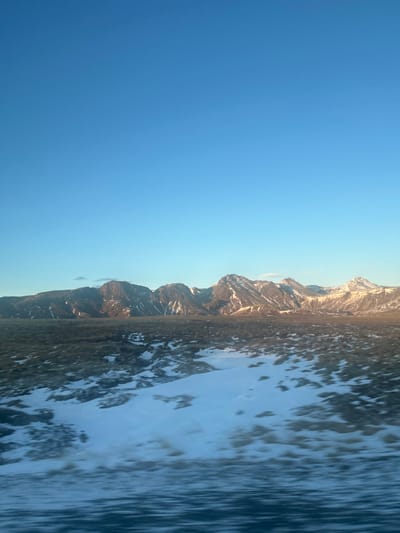 The image displays a vast, open winter landscape located in the vicinity of Laugarvatn, Iceland. The upper two-thirds of the frame are dominated by a clear, bright blue sky with a slight gradient, transitioning to a lighter hue near the horizon. A continuous range of rugged, dark mountains spans the middle ground, featuring exposed rock and significant snow cover on their slopes and peaks. Low-angle sunlight illuminates the rightmost peaks, highlighting their snow-capped surfaces with a warm, golden tone, while the mountains to the left are darker, possibly in shadow. The foreground and midground consist of a broad expanse of terrain covered by dark, low-lying ground vegetation or exposed earth, heavily interspersed with irregular patches of white snow. Horizontal motion blur is evident across the bottom portion of the foreground. No individuals, animals, or man-made structures are visible within the scene.