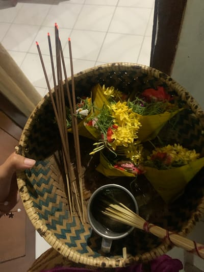 In a dimly lit interior, possibly a home in Sukasada, Indonesia, a woven bamboo basket is held by a person's left hand. The basket is filled with several small, cone-shaped offerings wrapped in yellow leaves, containing an assortment of colorful flowers, including yellow and red blooms, and green foliage. Standing upright within the basket are several thin incense sticks, their tips glowing red from being lit, emitting smoke. To the right of the incense sticks, a small metal cup or container is visible, holding a bundle of unlit incense sticks tied together with red string. The background appears to be a tiled floor and a wooden surface. The overall scene suggests a ritualistic or devotional practice.