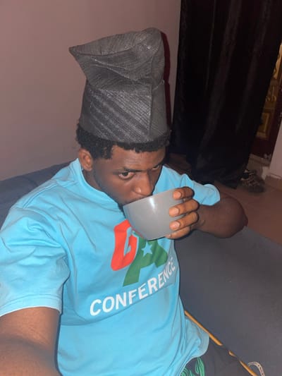 A single adult male is depicted indoors, framed from the mid-torso upwards. He has dark skin, short dark curly hair, and wears a dark grey, textured, folded cap. His attire includes a light blue short-sleeved t-shirt with a multi-colored "GA CONFERENCE" logo on the chest, featuring red, green, and white elements. The man is actively drinking from a plain grey bowl, which he holds with both hands, with his mouth positioned at the rim. His eyes are open and directed towards the camera. He is seated on a dark grey upholstered surface. The background features a pale, indistinct wall on the left, transitioning to a dark curtain on the right. Below the curtain, a light-colored tiled floor is visible, along with dark footwear. The scene is located in Ube, Nigeria.