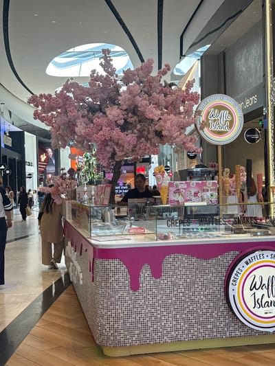 In the city of Leidschendam, Netherlands, within a modern shopping mall, a brightly decorated ice cream and waffle stand, "Waffle Island," is the focal point. A large artificial cherry blossom tree with delicate pink flowers is positioned to the left of the stand, adding a whimsical touch. A woman wearing a beige outfit stands behind the counter, appearing to be interacting with a customer. The stand features a display case filled with ice cream tubs and an assortment of waffle cones. Promotional posters showcasing menu items are visible on the stand. The surrounding environment includes shops with prominent signage, such as "Calvin Klein Jeans" and "Hollister." Other shoppers can be seen in the background, navigating the mall's polished floors and walkways. The scene captures a vibrant commercial setting within an indoor shopping complex.