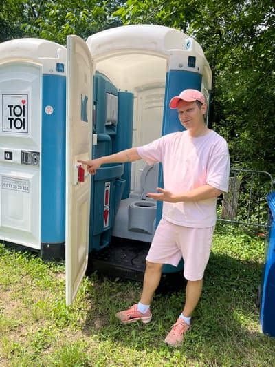 A young man stands outdoors next to an open blue and white portable toilet on a bright, sunny day. He is fair-skinned, wearing a light pink t-shirt, matching pink shorts, a pink baseball cap, white socks, and pink sneakers. He has a small tattoo on his left forearm. The man is smiling and looking directly at the camera, posing with his left hand gesturing towards the portable toilet's open door latch, and his right hand slightly extended.
The scene is set in a grassy area with lush green trees and foliage visible in the background, suggesting an outdoor event or recreational space. The bright sunlight casts clear shadows, indicating it is daytime. The portable toilet, an object often used at outdoor gatherings or construction sites, is a prominent feature.
Visible text on the side of the portable toilet includes the company name "ΤΟΙ ΤΟΙ d.o.o. Mobilni Sanitarni Sistemi" and an address "Bulevar Kralja Aleksandra 177, Beograd," along with a phone number "011 38 20 434." This text suggests the portable toilet belongs to a Serbian company, aligning with the provided location context of Sremski Karlovci Municipality, Serbia. The overall mood is casual and cheerful, with the man's pleasant expression contributing to the relaxed atmosphere.