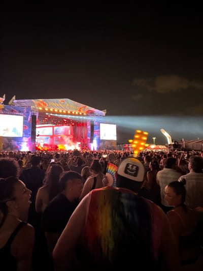 The image depicts a crowded outdoor music festival at night, likely in Rivas-Vaciamadrid, Spain, given the presence of a Johnnie Walker sponsored stage. The stage is brightly lit with red and white lights, featuring large screens on either side displaying abstract visuals or performances. A large crowd of people, seen from behind in the foreground, is gathered in front of the stage, many holding up their phones to capture the event. The sky above is dark with scattered clouds. In the mid-ground, a structure with many illuminated orange lights is visible, as well as a large white arch-like structure. The overall atmosphere is that of a vibrant concert or festival.