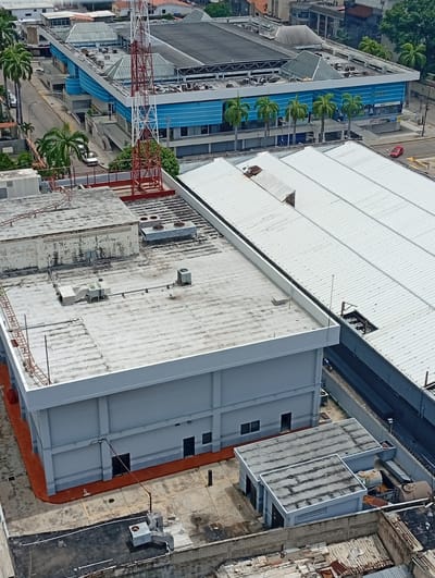 The image shows an aerial view of a commercial area in Valencia, Venezuela. In the foreground, a grey industrial building with orange-trimmed foundations has several windows and doors. Adjacent to it, on the same level, are two smaller grey structures with pitched roofs. Further back, a large building with a white, corrugated metal roof dominates the scene, with a blue-accented commercial building behind it. A prominent red and white communication tower is visible in the mid-ground. Palm trees line the street in front of the blue building. Several cars are parked on the street and a red car is visible on the road. The overall impression is of a modern, functional urban environment.
