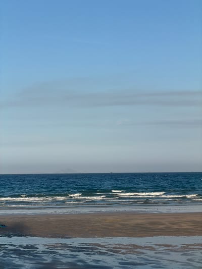 This is a vertical, medium-shot, eye-level photograph of a beach at Cam Lam, Vietnam. The image is divided into three horizontal sections: sky, sea, and shore. The sky is a pale blue with faint horizontal cloud formations. The sea is a deep blue, with several lines of white waves crashing on the shore. The shore consists of wet sand with tidal pools reflecting the sky and sea. In the distance, on the horizon, are a few faint shapes that appear to be ships or islands. In the lower left corner, a small portion of a blue and green object, possibly a kite or beach toy, is visible. The overall scene is serene and captures the natural beauty of the coastline.