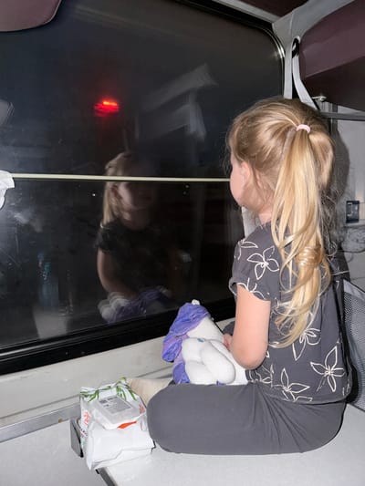 The image displays a young girl with long blonde hair, tied in a pink scrunchie, leaning forward and looking down into a dark, reflective surface, likely a window or table inside a vehicle. She wears a dark t-shirt adorned with a white floral pattern and dark grey or black pants. In her arms, she cradles a white stuffed animal, possibly a unicorn, partially covered by purple fabric.
The setting appears to be the interior of a vehicle, such as a train or bus, suggested by the visible seating on the left and the structure around the dark reflective panel. The dim lighting, with darkness outside, creates a prominent reflection of the girl's face and a distinct red light on the surface she's observing. A grey strap, resembling a seatbelt or luggage strap, is also visible.
To her right, on a narrow ledge, sits a pack of wipes or tissues. Visible Cyrillic text on the packaging includes "ДЛЯ ВСЕЙ СЕМЬИ" (For the whole family) and "ВЛАЖНЫЕ САЛФЕТКИ" (Wet wipes). The girl's posture suggests an engaged or curious demeanor as she observes her reflection and the outside darkness. The scene evokes a quiet moment during travel, set in Michurinsk, Russia, likely in the evening or at night.