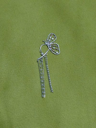 A silver butterfly-shaped earring with pink and clear rhinestones is positioned on a textured green fabric surface. The earring features delicate chain dangles adorned with small beads. The background is a uniform, soft green. The image is a close-up shot of the jewelry.