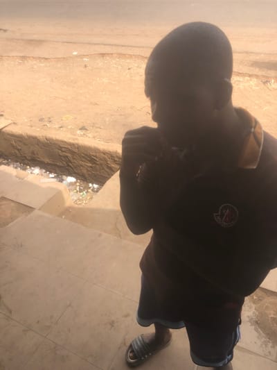 The image depicts an outdoor scene in Kufena, Nigeria, viewed from a slightly elevated perspective. A single male individual is positioned in the foreground, facing partially away from the camera and towards the left. His facial features are obscured by shadow, but his right hand is raised near his mouth or chin. He wears a dark short-sleeved polo shirt with a yellow-gold collar and a circular Moncler emblem on the left chest. Below, he wears dark shorts with a light blue stripe along the bottom edge, and light-colored slide sandals on his feet. The individual stands on a light-toned, tiled surface. Immediately beyond this surface, a low concrete curb defines the edge of a narrow ditch or gutter filled with miscellaneous refuse, including plastic containers and paper debris. Further in the background, a wide, unpaved, dusty road extends horizontally across the frame, exhibiting minor surface undulations. The scene is illuminated by bright outdoor light, casting the foreground subject into relative shadow.