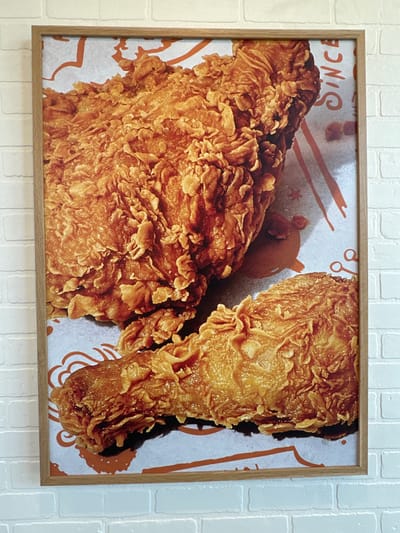 The image shows a framed poster of fried chicken hanging on a white brick wall. The poster features two pieces of crispy fried chicken, with one large piece in the upper left and a drumstick in the lower center. The background of the poster has orange graphic elements and text that reads "SINCE" vertically on the right side. This poster is located at Popeyes Louisiana Kitchen at 329 North Road, Coquitlam, BC, Canada.