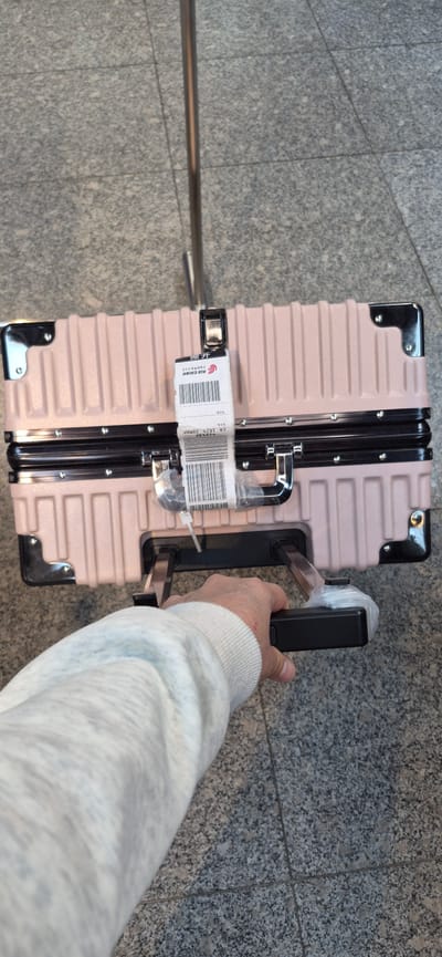 The image shows a person's hand and arm, wearing a light-colored sweater, holding the retracted handle of a light pink, hard-shelled suitcase. The suitcase has a ribbed texture and is adorned with metallic corner protectors and latches. A white baggage tag, featuring a barcode and text that includes "AIR CHINA" and "CA 1629 2DMAF", is attached to one of the suitcase's latches. The suitcase is placed on a gray, speckled floor, likely made of granite tiles, with visible grout lines. In the background, a metallic railing or pole runs horizontally across the frame. The scene appears to be indoors, possibly in an airport or a similar travel terminal, given the flooring and the baggage tag. The lighting suggests it is daytime.