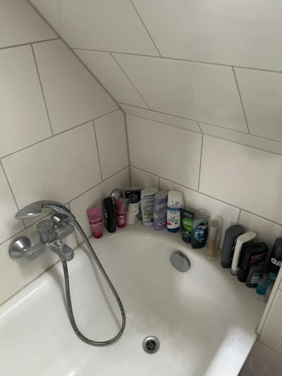 The image depicts the interior of a bathtub in a Gelsenkirchen, Germany residence. The bathtub is white and appears to be made of porcelain or enamel. Various toiletries, including shampoo bottles, body wash, and lotions, are arranged along the edge of the tub. The walls surrounding the bathtub are tiled with white square tiles, with grey grout lines creating a geometric pattern. A chrome faucet and shower head are attached to the wall above the tub. The lighting suggests an indoor, possibly domestic, setting.