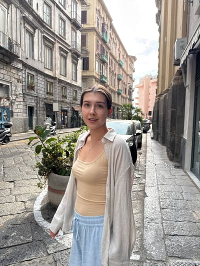 The media file shows a young woman standing on a cobblestone street in what appears to be Naples, Italy. She is looking towards the camera with a slight smile. The woman is wearing a light-colored, possibly linen, open-button shirt over a beige tank top and light blue pants. The background features old, multi-story buildings with numerous windows and balconies, characteristic of Mediterranean architecture. A potted plant is visible next to her, and a parked motorcycle is in the lower left corner. The street is paved with cobblestones, and there are some cars parked further down the street. The lighting suggests it is daytime, possibly a sunny or overcast day. The overall mood appears relaxed and casual. No visible text is present in the image.