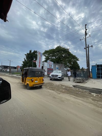 The image captures a street scene in Epe, Nigeria, under a cloudy sky. A yellow auto-rickshaw (tricycle) is prominently positioned in the lower center of the frame, moving towards the right. Behind the auto-rickshaw, a white Mercedes-Benz SUV is parked on the side of the road. A man dressed in a white shirt and trousers stands near the SUV. Street signage is visible behind the vehicles, including a sign for 'Foursquare.' Overhead are electrical lines supported by wooden poles. The road surface appears to be made of concrete or asphalt, exhibiting signs of wear and tear. The foreground contains a partial view of the vehicle from which the photo was taken.