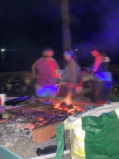 In Marbella, Spain, at night, three individuals are gathered around a bonfire, with a grill placed over the embers. The scene appears to be outdoors, possibly near a beach or waterfront, as indicated by a distant light on the water. The fire is actively burning, casting a warm glow on the surrounding area and reflecting off the participants. Two individuals are standing, while the third is partially obscured. The atmosphere is illuminated by the bonfire and some colored lights, likely from artificial sources, creating a dynamic and somewhat blurred effect due to the low light conditions. A palm tree trunk is visible in the background. The ground is covered with gravel and ash from the fire.