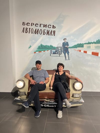 In the Verkhnyaya Pyshma, Russia, the image depicts a man and a woman posing on a prop car. The car, a vintage model with a beige exterior and brown interior, is positioned in front of a mural. The mural features a street scene with a white and orange checkered barrier, a building, and trees. A figure in a blue uniform, possibly a police officer, stands near a motorcycle in the mural.

The woman, wearing a black tank top and black pants, reclines on the car's seat, her arm resting on the headlight. She has dark hair and smiles at the camera. She carries a black bag slung across her body. The man, dressed in a grey t-shirt, black pants, and a black baseball cap, sits on the other side of the car, looking towards the camera with a smile. He wears a watch on his left wrist. Both are wearing athletic shoes.

The backdrop is a white wall with the mural. On the left side of the mural, text is visible in large, dark brown Cyrillic letters: "Берегись АВТОМОБИЛЯ" which translates to "Beware of the car".

The scene appears to be indoors, possibly at a museum or exhibition, given the presence of the prop car and the stylized mural. The lighting suggests an indoor environment, with no indication of natural light or weather conditions. The overall mood is playful and lighthearted, with both individuals smiling and posing for the photograph.