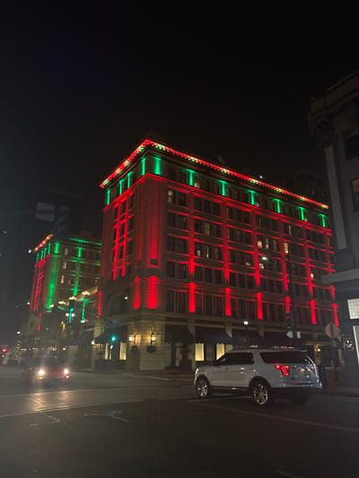 A tall building is illuminated with green and red lights, reminiscent of Christmas decorations. The building is multi-storied, with a visible pattern of windows. A white vehicle is parked on the right side of the frame. The scene takes place at night, and the overall lighting is subdued, accentuating the building's colorful facade.