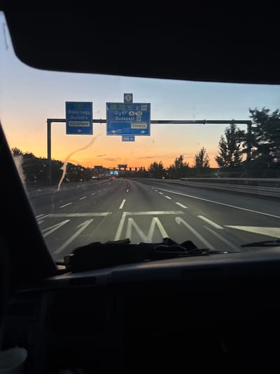 The image is taken from inside a vehicle, looking out through the windshield at a highway scene during sunset. Overhead highway signs indicate directions to "Biatorbágy", "Budaörs", "BUDAPARK", "Győr", "Budapest", and "TOPARK". One sign also shows "M1" and "M7". The sky is a gradient of orange, pink, and blue, indicating the sun is low on the horizon. Trees line the sides of the highway, and other vehicles are visible in the distance. The dashboard and part of the vehicle's interior are visible in the foreground. The scene appears to be on a major highway leading into or out of Budapest, possibly near the city of Biatorbágy, Hungary, given the signage.