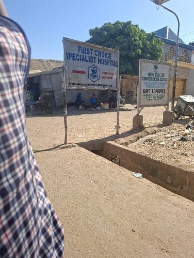 This image depicts an outdoor street scene in Dan Maliki, Nigeria, under a clear blue sky. In the immediate foreground on the left, a portion of a person's torso, clad in a blue, white, and red plaid garment, is visible. The foreground consists of a light-colored concrete or paved surface, which transitions into an open, concrete-lined drainage ditch running horizontally across the mid-ground. A discarded plastic bottle and other refuse are present within the ditch.
In the middle ground, two weathered freestanding signs are prominent. The left sign, primarily white with blue text, reads "FIRST CHOICE SPECIALIST HOSPITAL" and features a red directional arrow pointing left, a logo, and contact information including phone numbers (08030097123, 09033329283) and an email address (firstchoicespecialisthospital@gmail.com). Beneath this sign, several individuals are gathered, approximately 4-5 people, seated on low stools or the ground, some with containers, suggesting an informal market or gathering. One individual wears a blue garment. Behind them are simple, single-story structures with corrugated metal roofs. A motorcycle is partially visible behind the left sign.
The right sign, also white with black text, advertises "HIGH QUALITY COMPREHENSIVE SCHOOLS PRE-NURSERY, NURSERY, PRIMARY & COLLEGE GOVT. APPROVED," followed by additional text related to management and contact numbers (08036987342, 08139414909). To the right of this sign, a two-wheeled utility cart is visible.
In the background, a large, dense green tree stands to the right of the hospital sign. Further back, buildings with varied architectural styles, including one with a blue-tiled roof, are visible under the clear sky. A metal utility pole with a small solar panel extends from the right side towards the sky. Strong sunlight casts distinct shadows across the scene.