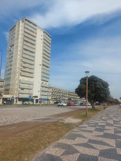 The image captures a bright daytime scene in Figueira da Foz, Portugal, under a clear blue sky with sparse white, wispy clouds. The foreground and right side of the frame are dominated by a distinctive patterned mosaic pavement, characteristic of "calçada portuguesa," featuring interlocking geometric shapes in light and dark grey tones. This paving extends into the background, bordering a strip of dry, light brown grass, which in turn gives way to a road with parked cars.
On the left, a tall, slender, light-colored multi-story apartment building stands prominently, characterized by numerous horizontal balconies creating a stepped facade. In the mid-ground, a large, dense tree with a rounded green canopy partially obscures a modern streetlamp, which has a grey bell-shaped head and a brown pole. Behind the tree and across the road, another multi-story building with numerous balconies is visible, and more cars are parked along this street.
Several road signs and advertisements are discernible. A blue rectangular sign clearly displays "Gest. de Imóveis" (Real Estate Management). Another blue sign below it is partially obscured but starts with "Guesthouse da R...". Other visible signs include multiple blue square pedestrian crossing signs with white figures, a circular red 'no entry' sign, and a blue circular 'turn right' sign. Small, indistinct figures of people can be seen near the buildings and parked vehicles. The scene presents a typical, sunny urban street environment.