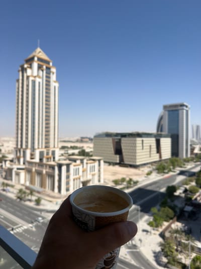 This vertical image presents a high-angle view of a modern cityscape, likely captured from a balcony or a tall building in Lusail, Qatar. In the foreground, a hand holds a white paper cup filled with a light brown liquid, presumably coffee, topped with a layer of foam. The cup itself features an intricate, dark-patterned design.
The background unfolds into an expansive urban landscape under a clear, bright blue sky, indicating daytime and sunny weather. Prominent in the scene are several contemporary high-rise buildings. To the left, a striking skyscraper with a distinctive golden-hued, pyramidal or pointed roof and white cladding stands tall. Further into the city, a large, rectangular building with textured facades and other modern structures are visible. Roads weave through the urban environment, and scattered patches of green foliage suggest landscaping or trees. While the overall impression is one of a bustling, developing city, the elevated perspective offers a moment of quiet contemplation with a beverage. No discernible text is visible on the cup or within the city view.