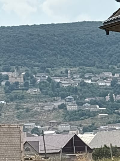A wide shot captures a hillside village with buildings nestled amongst dense green foliage and trees under a hazy sky. The village, identified as being in Ullubiyaul, Russia, features a mix of architectural styles, from older stone structures to more modern residential buildings with distinct rooflines. In the foreground, the roofs of several structures are visible, including one with a prominent triangular dark roof. A tall, thin pole stands vertically to the right of this structure. The hillside is terraced in some areas, suggesting agricultural activity. The overall impression is one of a rural, somewhat isolated community. No people or specific actions are discernible in the image.