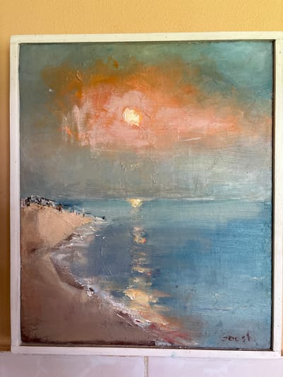 This is an oil painting of a seascape with a beach in the foreground. The sky is a dramatic mix of orange and blue, with a bright sun peeking through clouds. The ocean is a deep blue, reflecting the colors of the sky. The beach is sandy, with some people walking along it. There is a white frame around the painting. The signature on the lower right appears to be "Sgost" in dark red. There is no indication in the image or the provided information that this painting depicts or is located in Navapolatsk, Belarus.